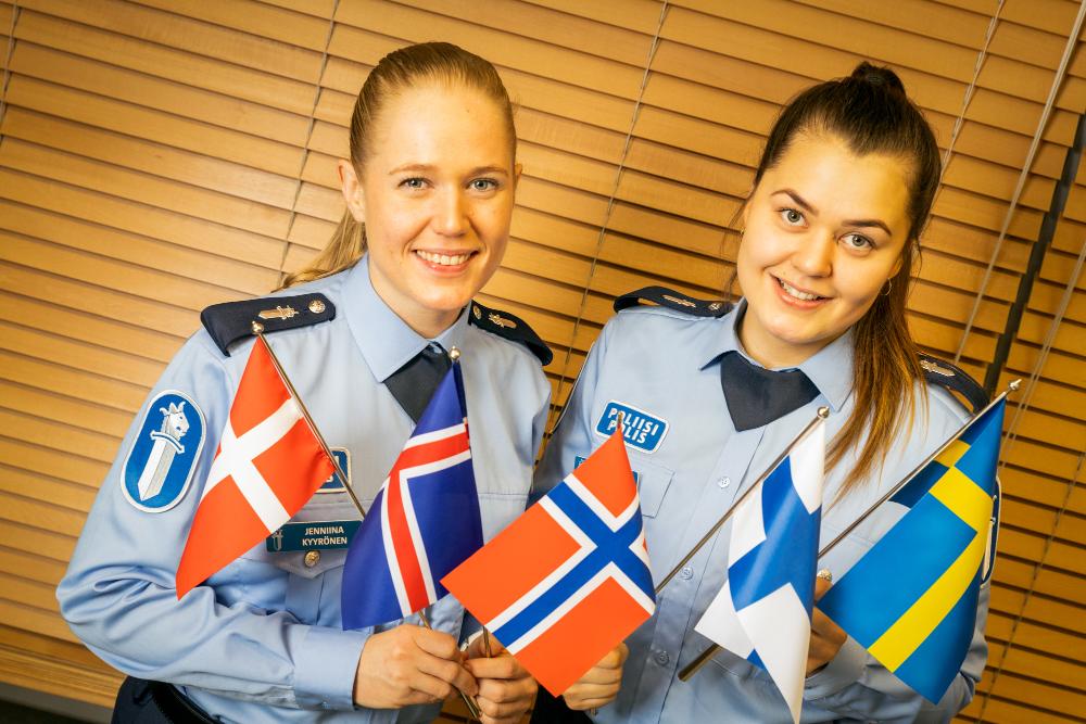 Kaksi hymyilevää poliisiopiskelijaa käsissään Pohjoismaiden pienoislippuja.