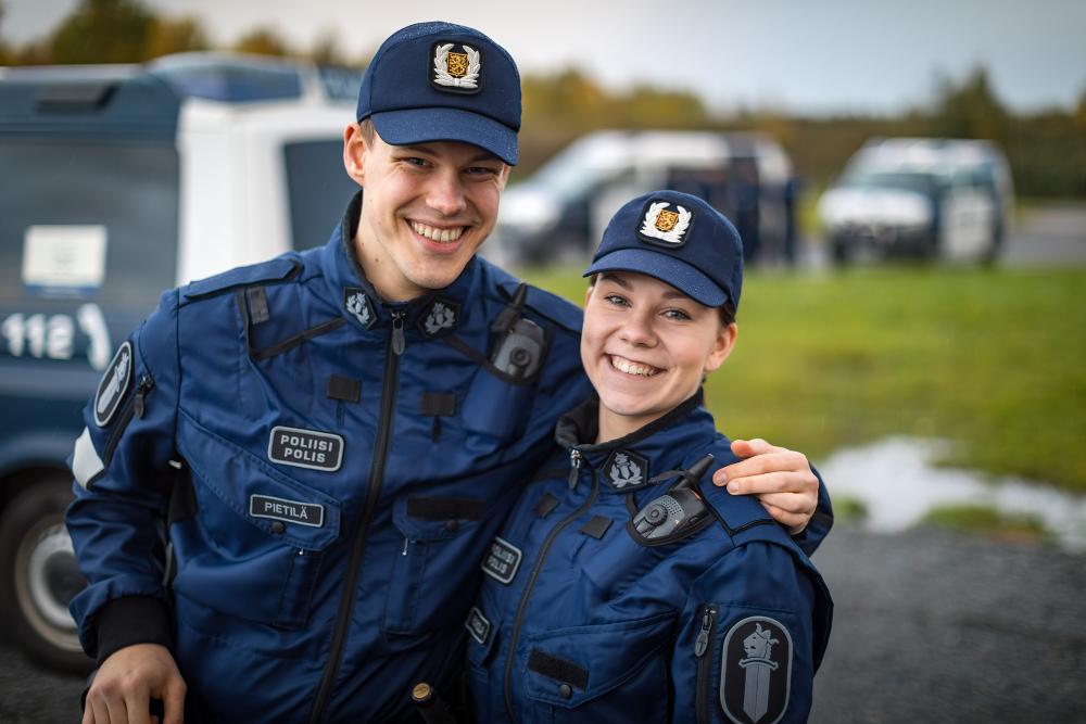 Two smiling police students in uniform with police cars behind them.